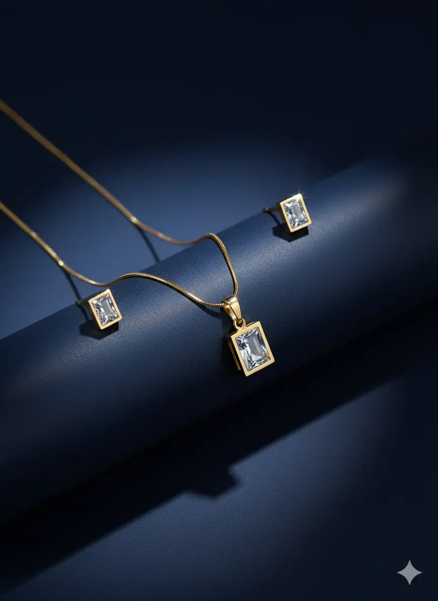 Minimal gold necklace with rectangular crystal pendant and matching stud earrings on dark background.