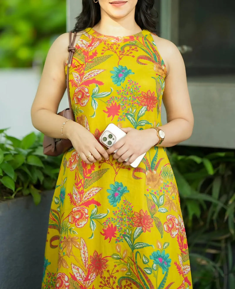 Sleeveless yellow dress with colorful floral print and a fitted waist, worn with a brown shoulder bag and white bracelet.