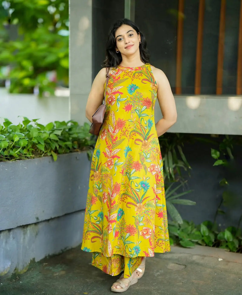 Yellow sleeveless dress with colorful floral and leaf print, flowy high-low hem, worn with sandals.
