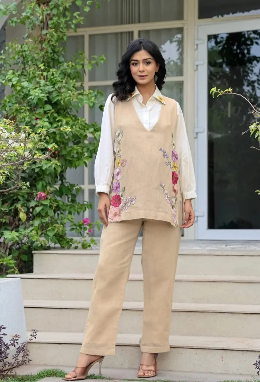 Beige knitted sleeveless corset with floral embroidery paired with matching pants and a white blouse.