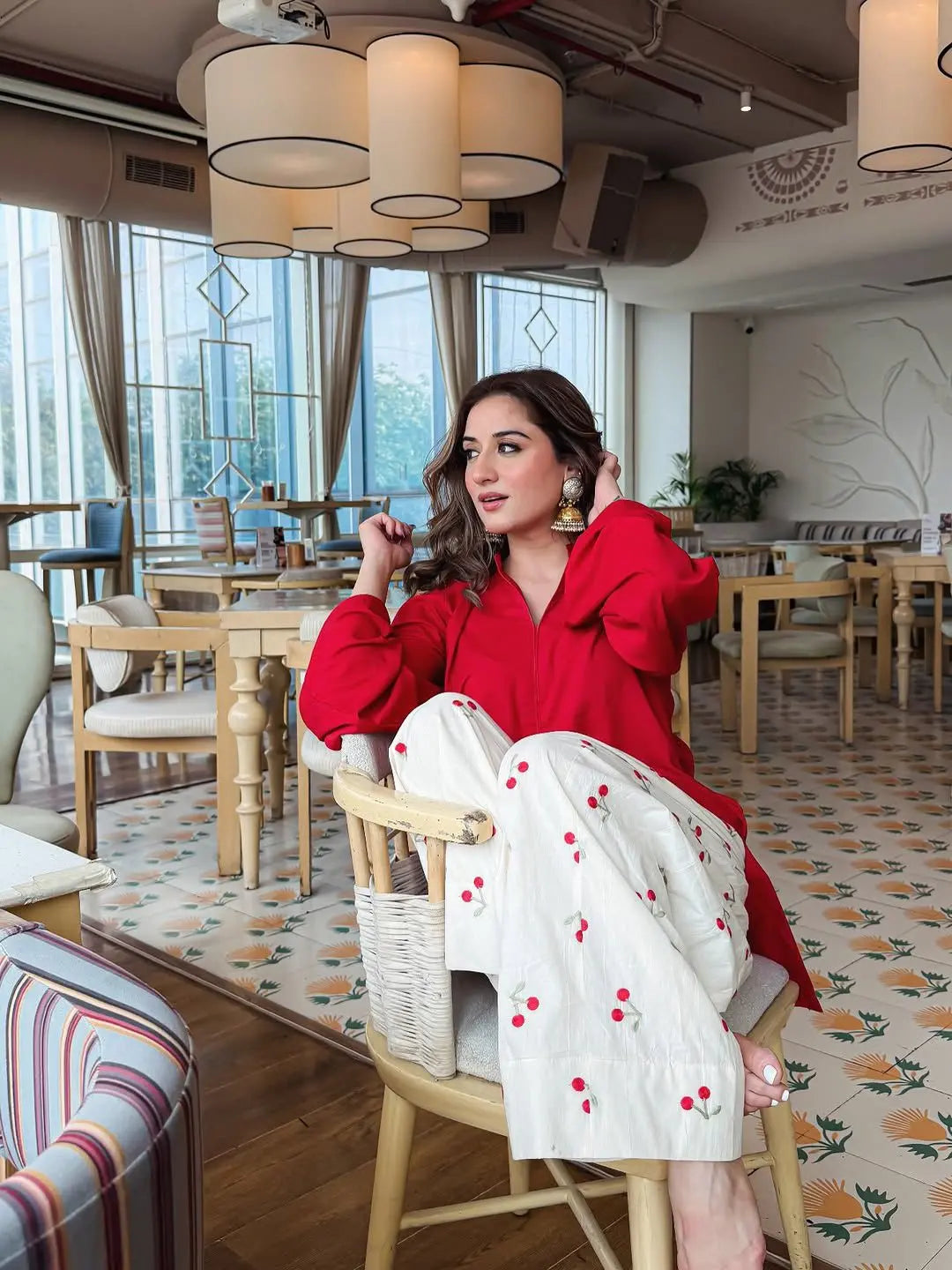 Woman wearing a red corset top with puffed sleeves paired with white pants featuring red cherry prints, seated in a modern.