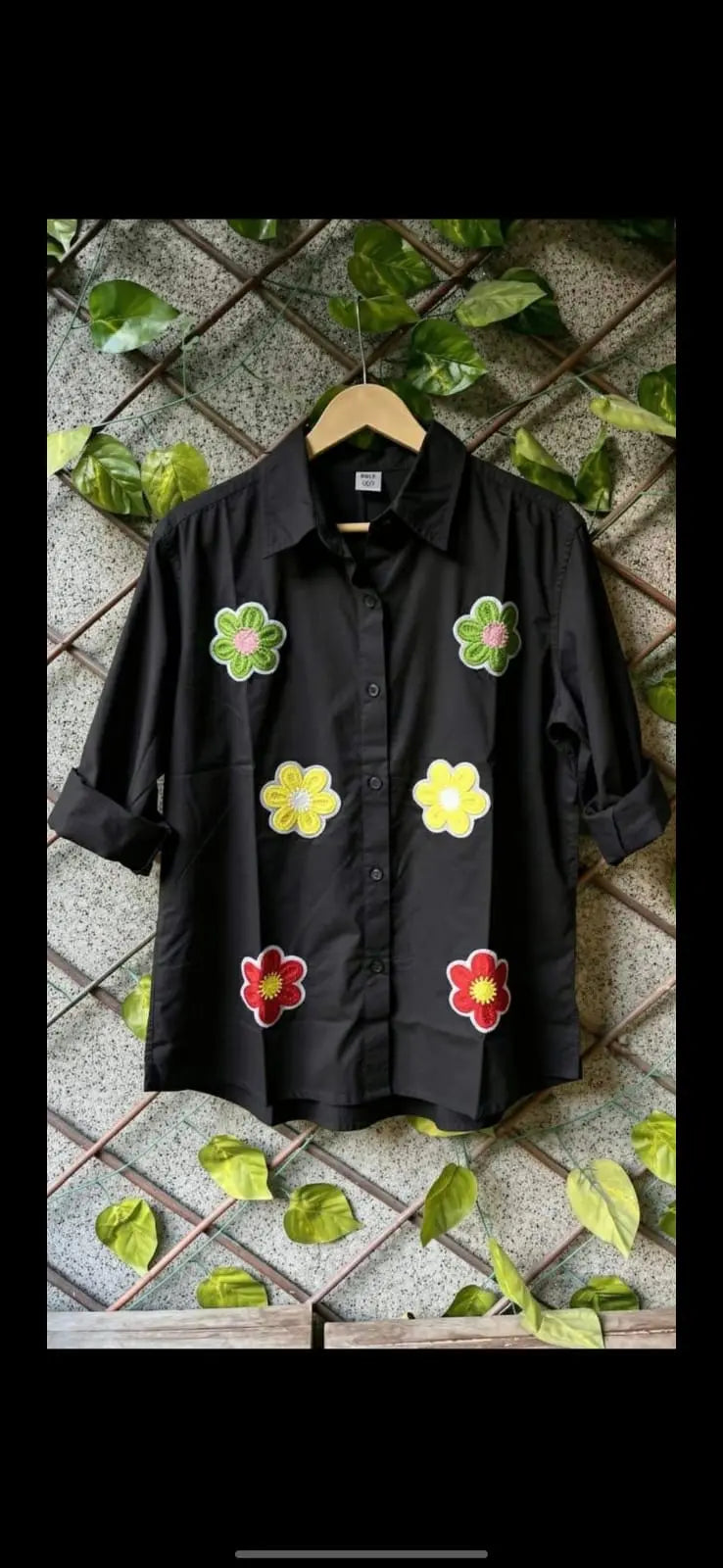 Black cotton shirt with colorful floral embroidery and rolled-up sleeves on wooden hanger.