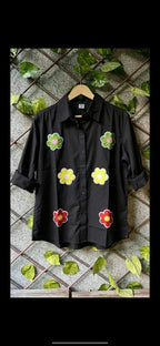 Black cotton shirt with colorful floral embroidery and rolled-up sleeves on wooden hanger.