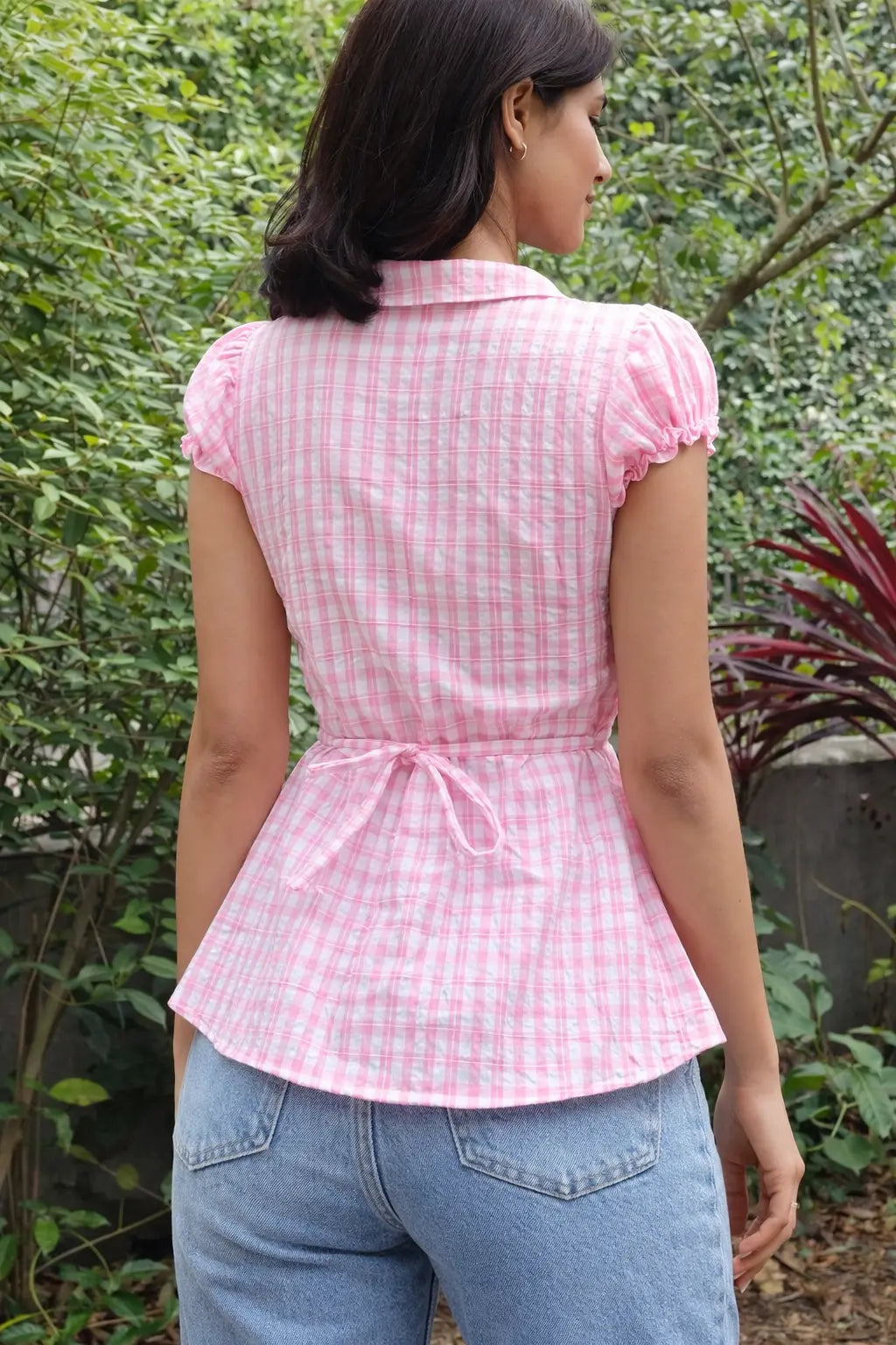 Woman wearing pink and white gingham peplum top with puff sleeves and a tied waist bow, paired with light blue jeans.